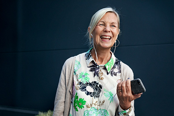 Eldre, smilende kvinne, med ørepropper, og iført fargerik overdel, holder en mobiltelefon og smiler.