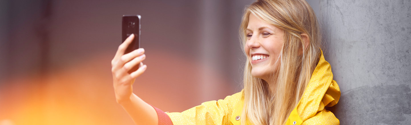 Blond kvinne i gul regnjakke smiler og tar selfie med mobilen mens hun lener seg mot en grå betongvegg.