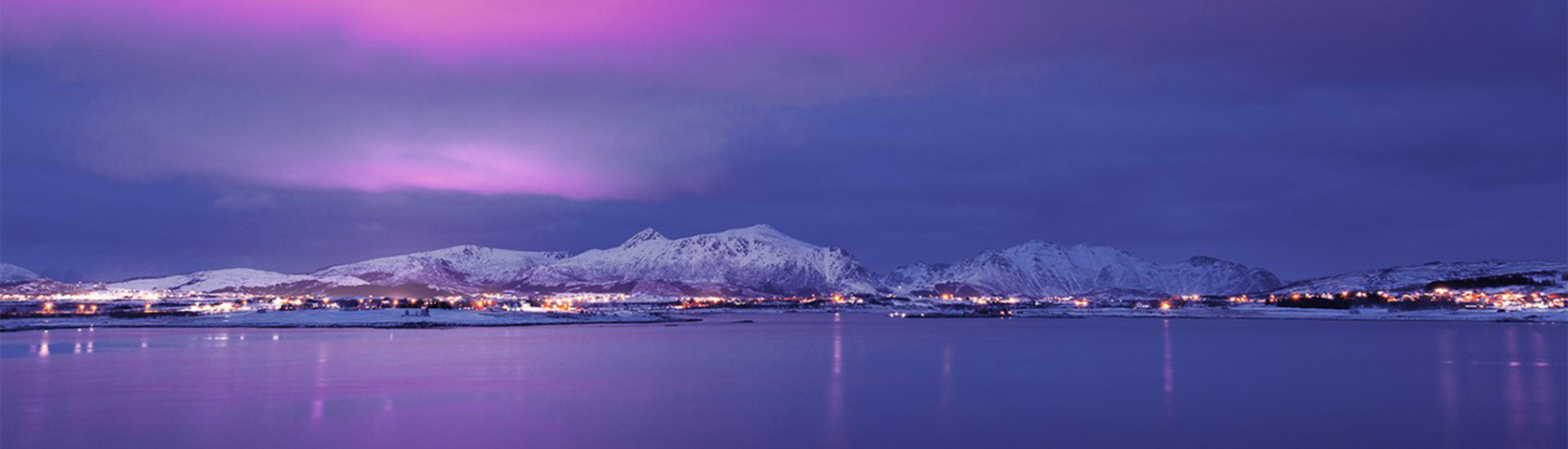 Utsikt over vinterkledd by som speiler seg i fjorden på kvelden.