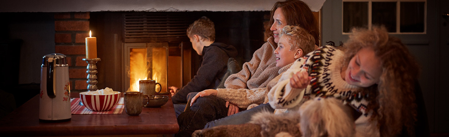 Familie sitter i sofaen på hytta. I bakgrunnen brenner det i peisen, og det er levende lys på bordet.