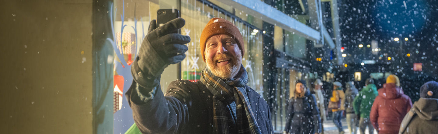 Eldre, smilende mann står og tar selfie ute i gata en snøfull kveld. Mange folk er ute, vi ser lys fra pyntede butikkvinduer, og julestemningen er til å ta og føle på.