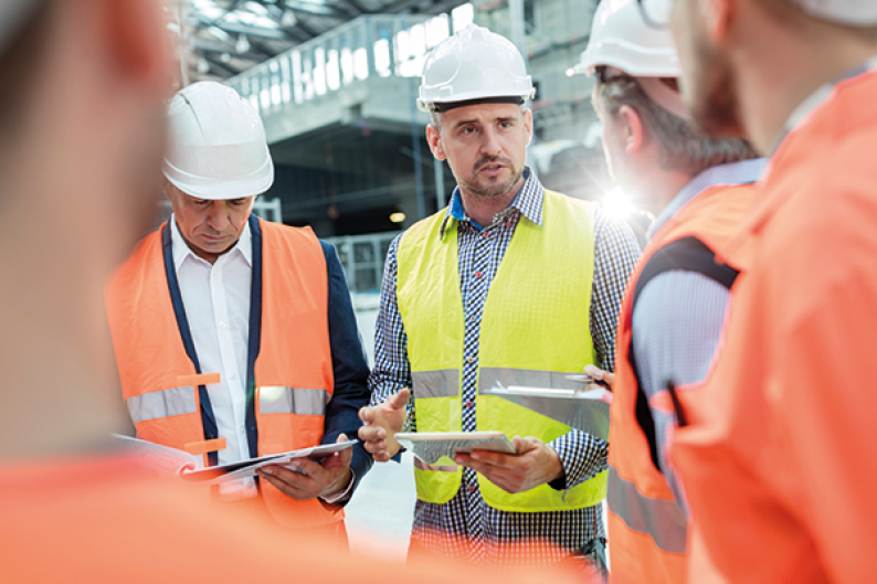 Arbeidskarer i gule / oransje vester og hjelm står og diskuterer noe.