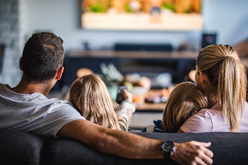 Familie sett bakfra sitter i sofaen og ser TV.