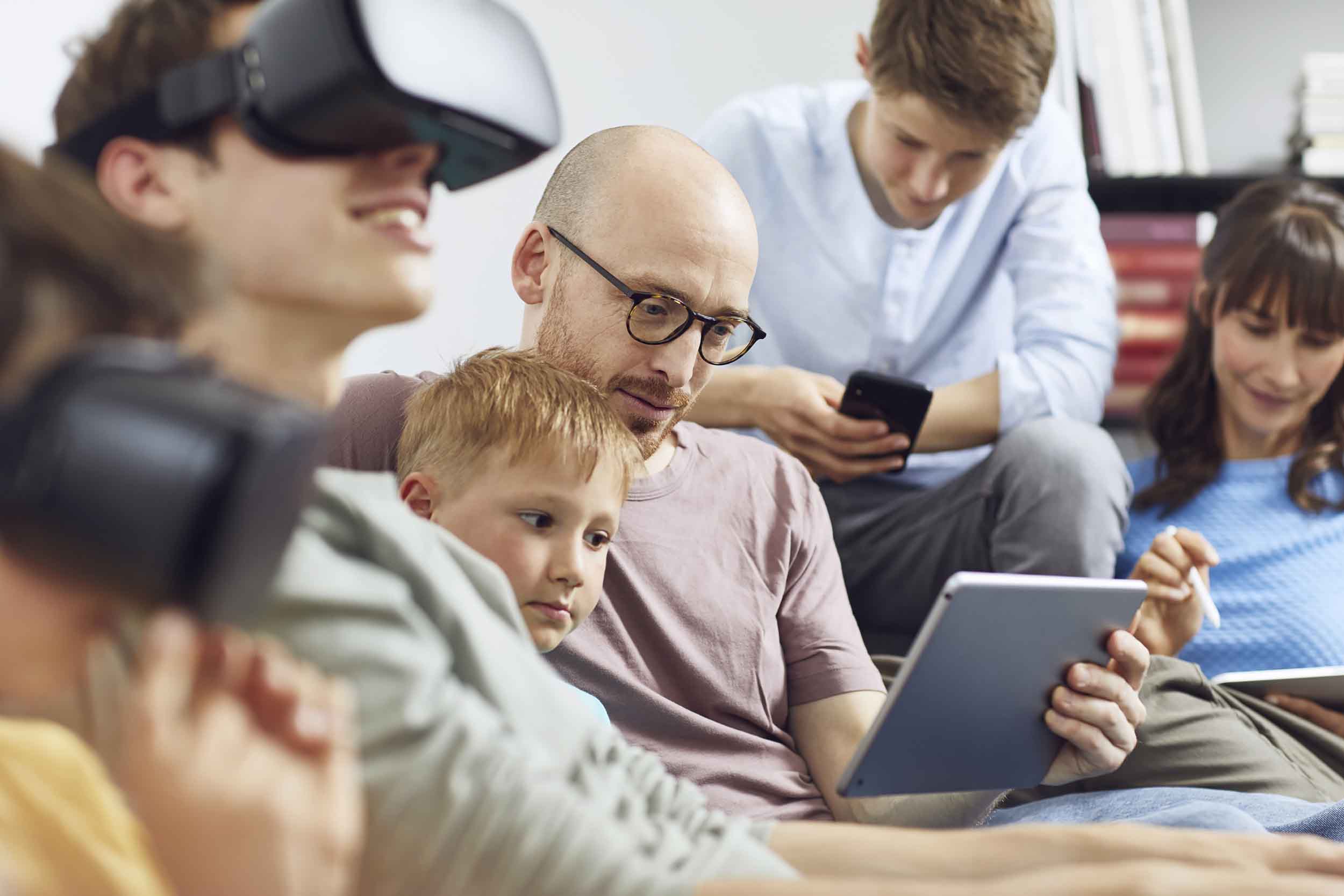 Bilde av en familie på 6 som sitter i en sofa. To stykker har på seg VR-briller, to sitter på mobilen, og to stykker ser på et nettbrett. Bilde av en familie på 6 som sitter i en sofa. To stykker har på seg VR-briller, to sitter på mobilen, og to stykker ser på et nettbrett.