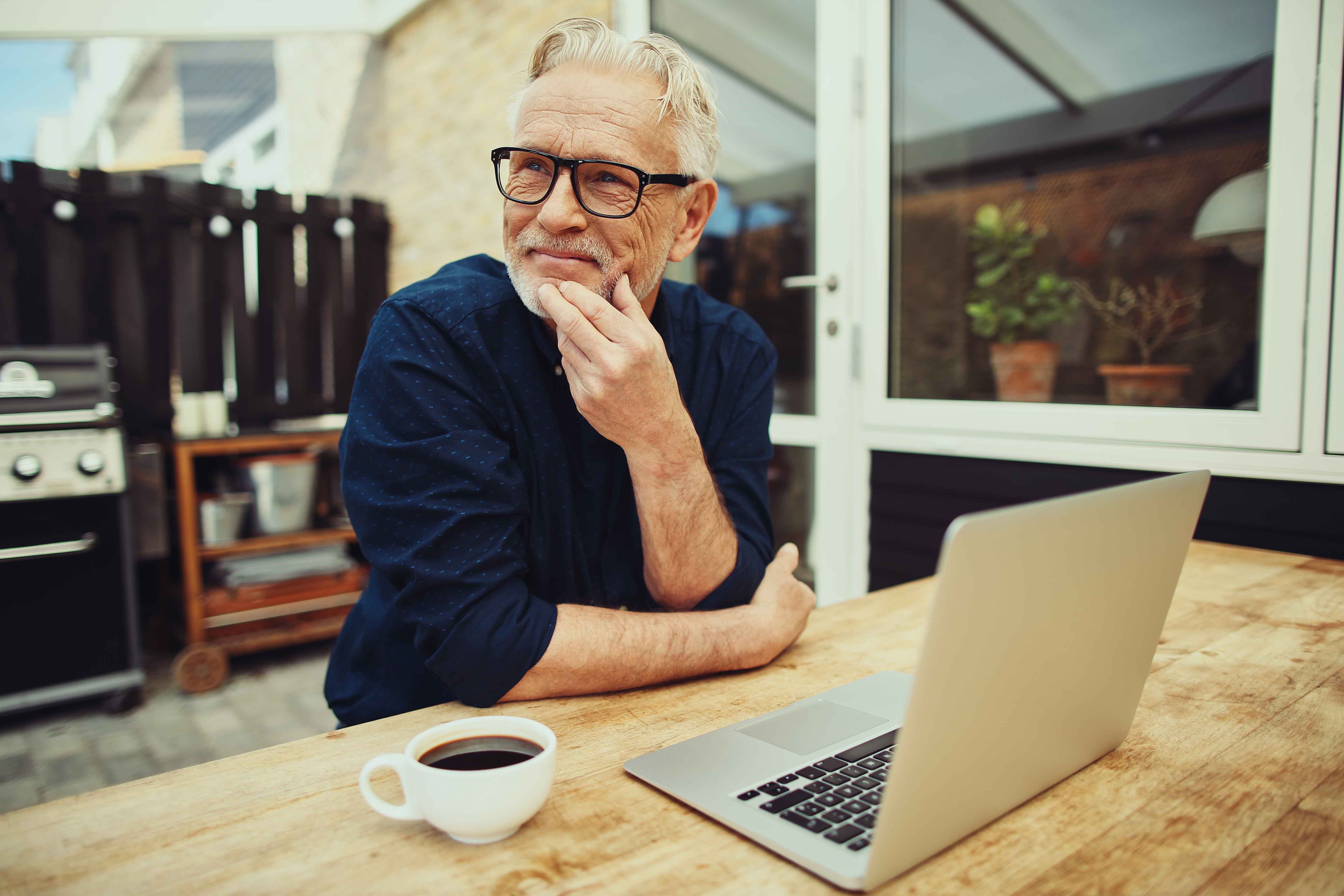 Bilde av en mann som sitter ved et bord ute på terrassen. Han har en PC og en kaffekopp på bordet Bilde av en mann som sitter ved et bord ute på terrassen. Han har en PC og en kaffekopp på bordet