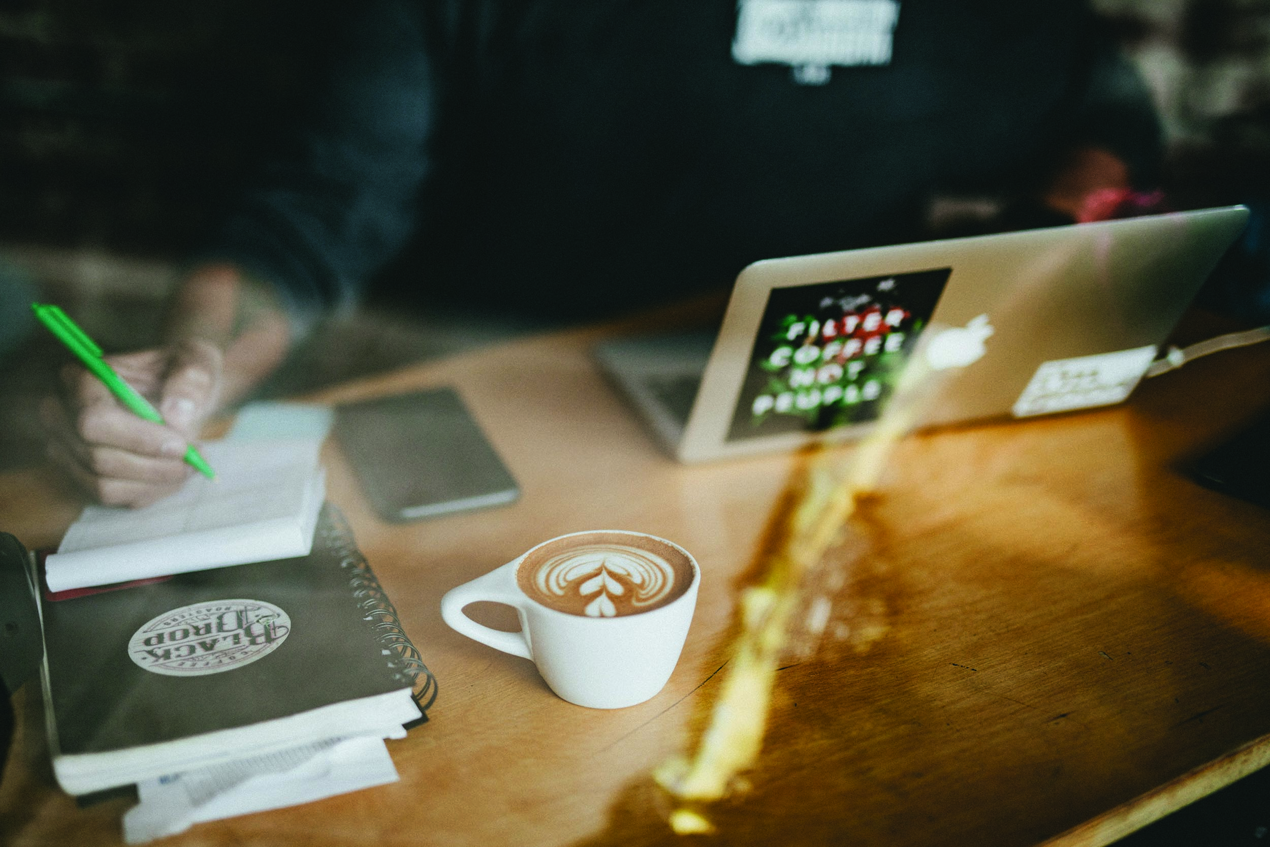 Bilde av et arbeidsbord. Det ligger en PC, en kaffekopp og noen notatbøker på bordet. Vi ser en hånd som skriver på en av bøkene. Bilde av et arbeidsbord. Det ligger en PC, en kaffekopp og noen notatbøker på bordet. Vi ser en hånd som skriver på en av bøkene.
