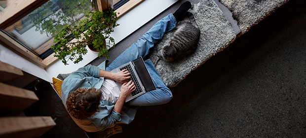 En mann og en katt sitter i en vinduskarm, mannen ser på en laptop