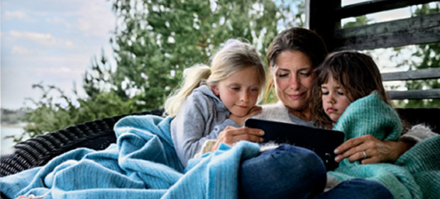 En liten familie sitter på en benk ute under et teppe og ser på et nettbrett