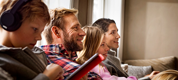 En familie sitter i en sofa og ser på en TV, mens en ung gutt ser ned på en telefon og har på seg høretelefoner.