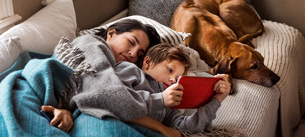 En mor og et barn slapper av på en sofa med en ipad og en hund sovende ved siden av.